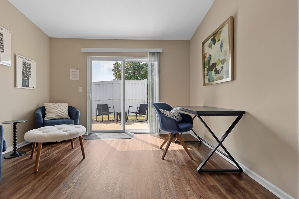 A small room with wood flooring, a desk and chair, an armchair with an ottoman, abstract wall art, and a sliding glass door leading to a patio with outdoor chairs.