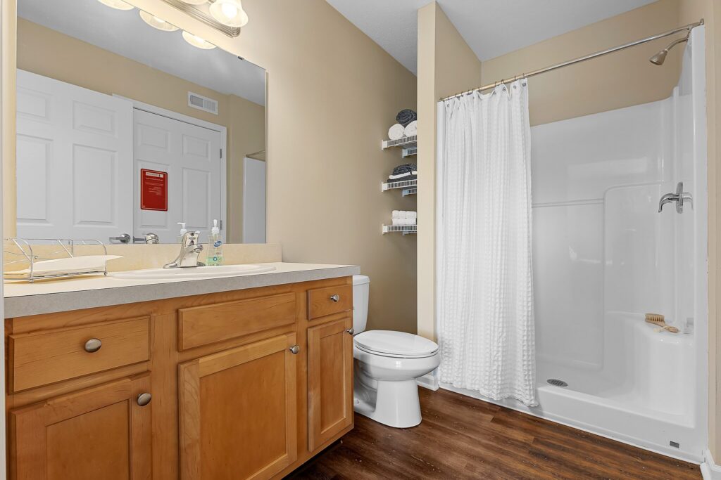 Modern bathroom with wood vanity, white countertop, toilet, shelves with towels, and a walk-in shower with a white curtain; tan walls and wood-look flooring.