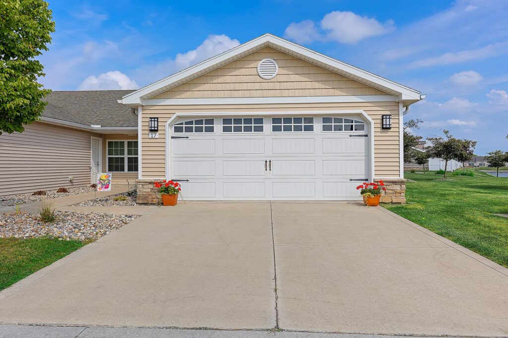 Single-story house with tan siding, white double garage door, concrete driveway, two potted flowers, and a small landscaped garden beside a trimmed lawn under a blue sky.