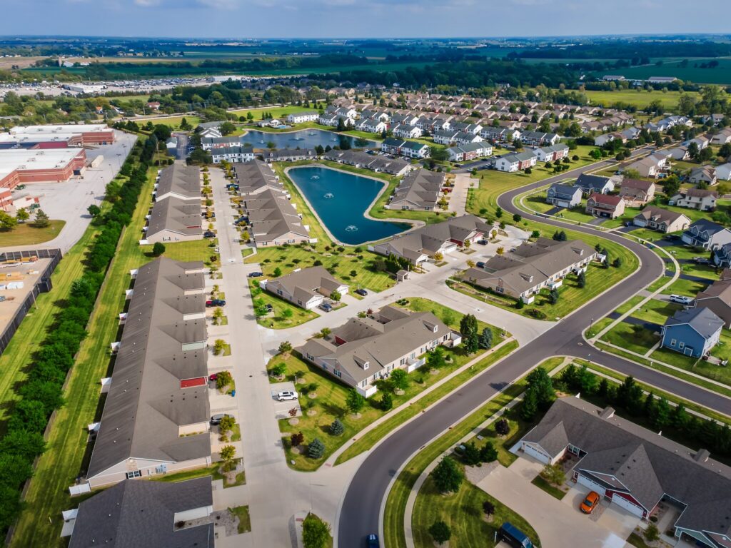 Aerial view of a suburban neighborhood with rows of houses, curved streets, green lawns, and a small pond at the center, surrounded by farmland and commercial buildings.