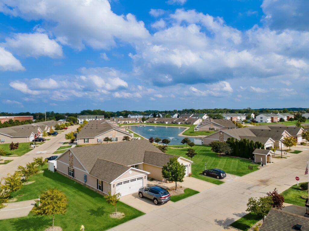 Aerial view of a suburban neighborhood with single-story houses, green lawns, parked cars, and a pond under a partly cloudy sky.