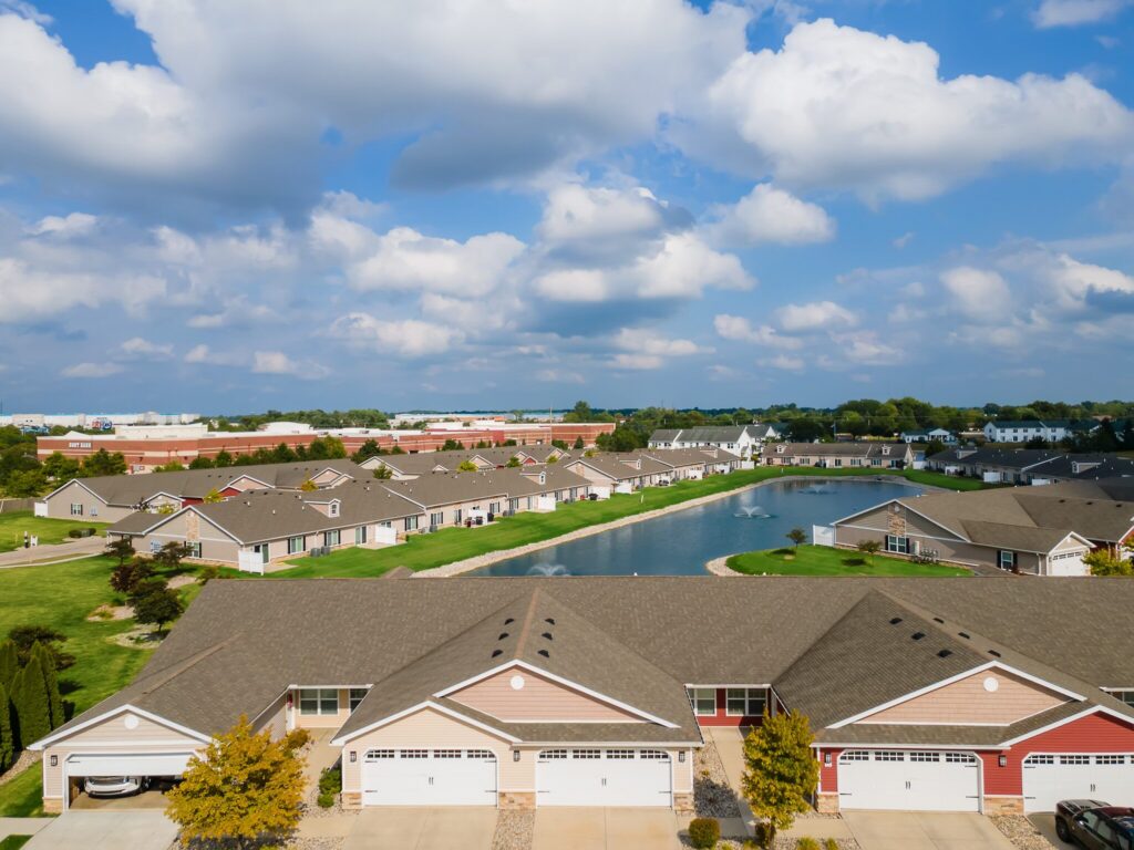 Aerial view of a suburban neighborhood with rows of attached houses, manicured lawns, a pond, and partly cloudy sky.