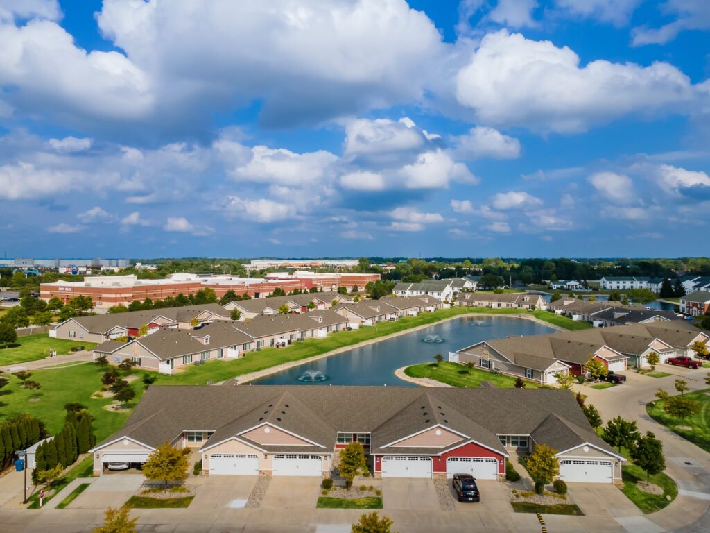 Aerial view of a suburban neighborhood with single-story houses, driveways, a pond with fountains, green lawns, and a partly cloudy sky.