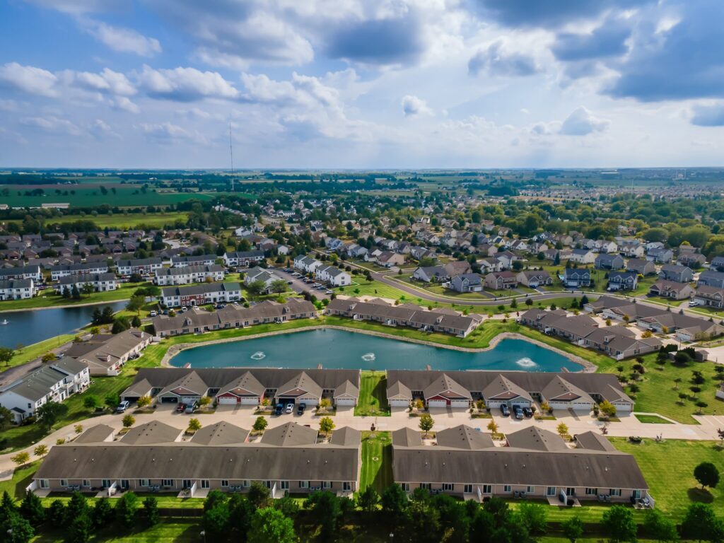Aerial view of a suburban neighborhood with rows of houses, small lakes with fountains, green lawns, and cloudy sky above.