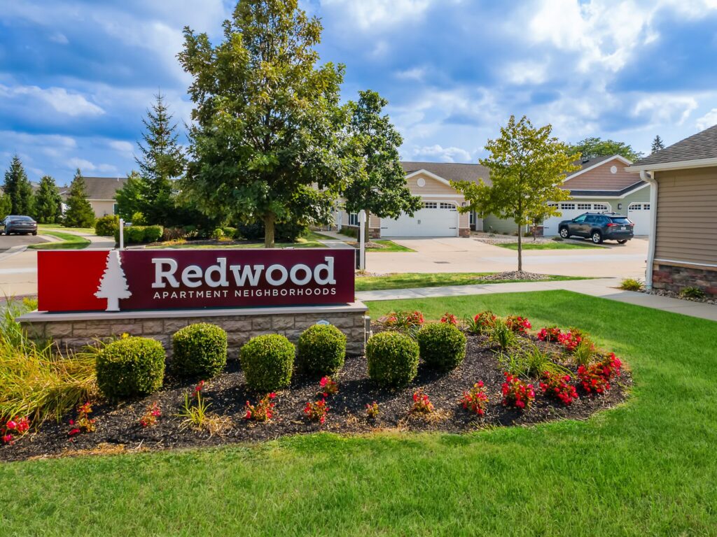 A sign for Redwood Apartment Neighborhoods stands in front of manicured landscaping with bushes and flowers; single-story homes and parked cars are visible in the background.