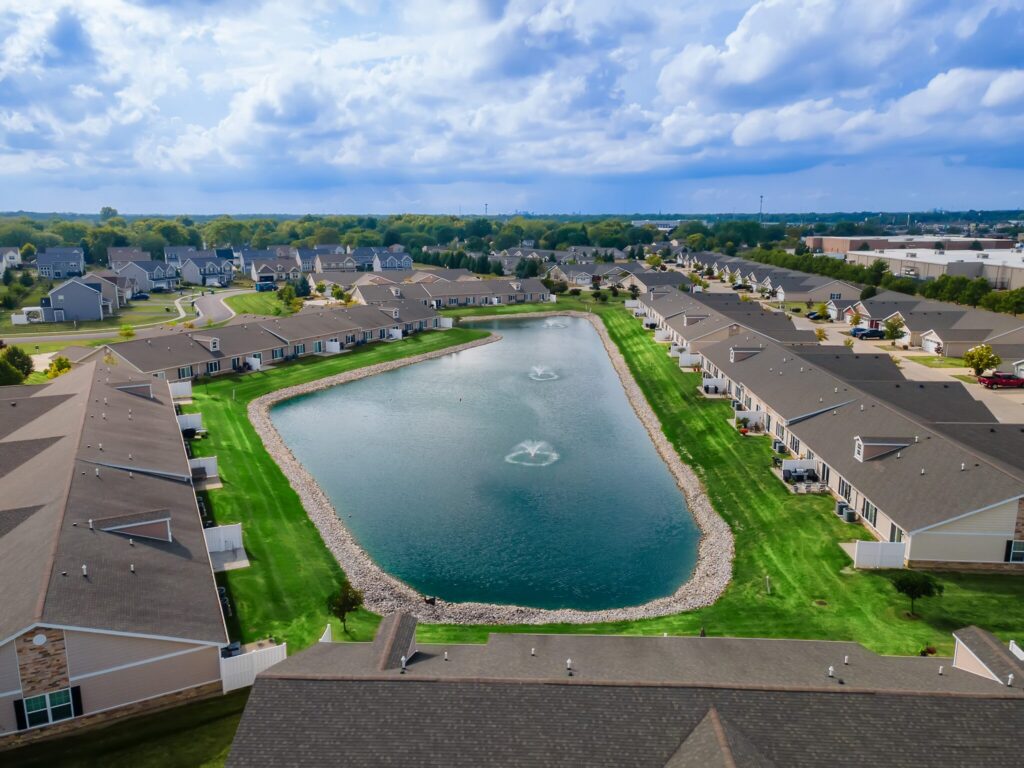 Aerial view of a suburban neighborhood with rows of houses surrounding a man-made pond with fountains, green lawns, and cloudy sky overhead.