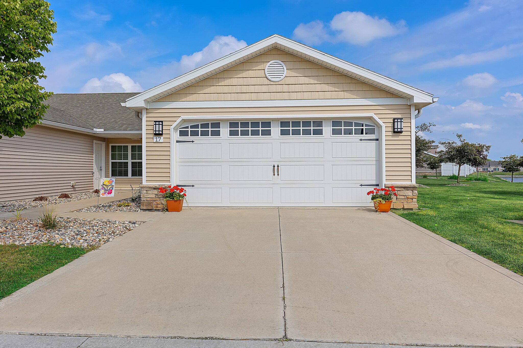 Single-story house with tan siding, white double garage door, concrete driveway, two potted flowers, and a small landscaped garden beside a trimmed lawn under a blue sky.