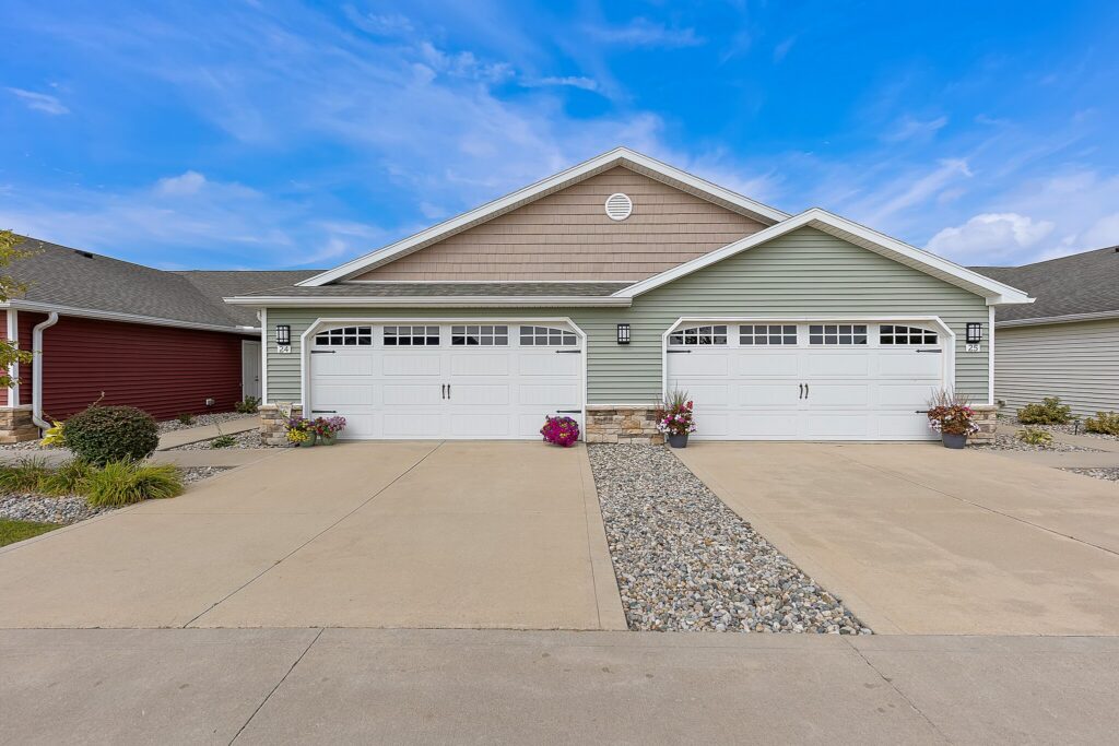 A duplex with two attached garages, stone landscaping, and potted flowers, set under a blue sky with scattered clouds.