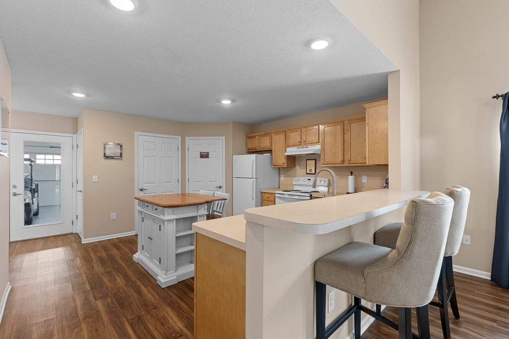 Modern kitchen with light wood cabinets, white appliances, a small island, and a breakfast bar with two upholstered stools. Hardwood flooring throughout the space.