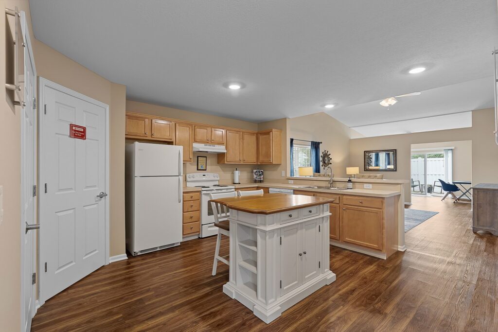 Modern kitchen with wooden cabinets, white appliances, a central island, and hardwood floors, opening into a bright living area with large windows and a ceiling fan.
