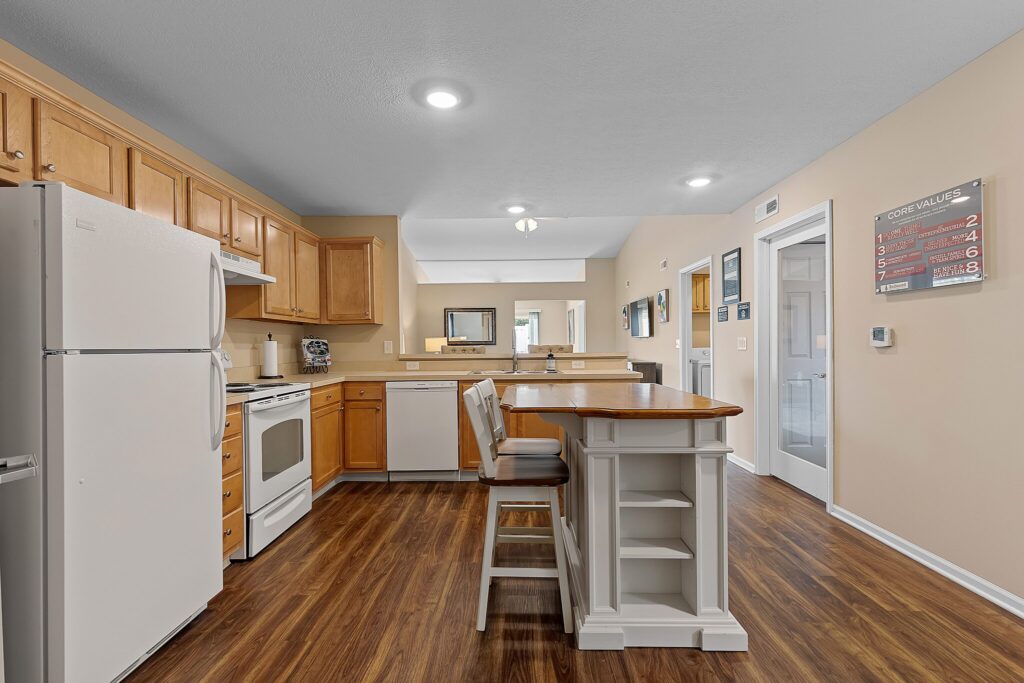 A kitchen with wood cabinets, white appliances, a central island with two stools, wood flooring, and beige walls with wall art and a thermostat.