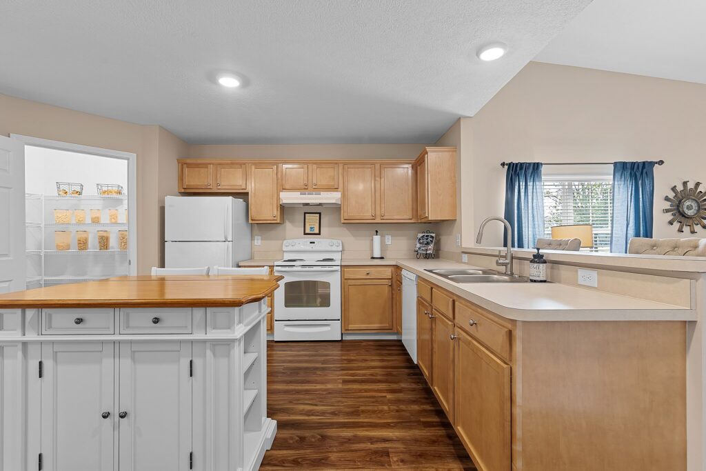 Modern kitchen with light wood cabinets, white appliances, a white island, beige countertops, and a window with blue curtains overlooking a dining area.