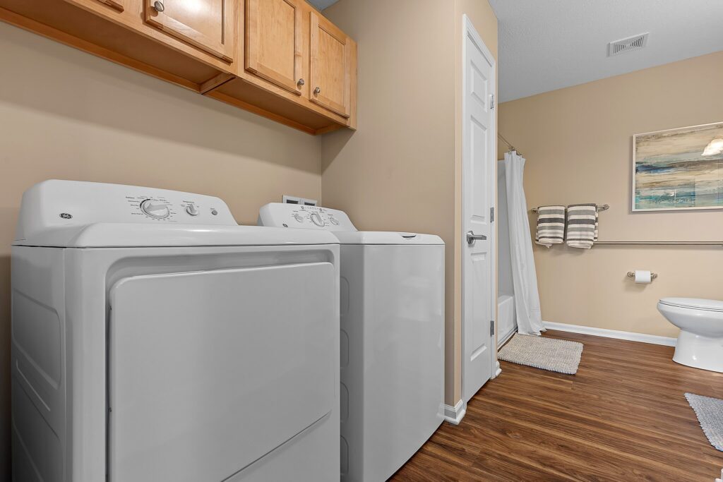 A laundry room with a white washer and dryer, wooden cabinets above, and a bathroom area with a toilet, shower, towel rack, and wall art.