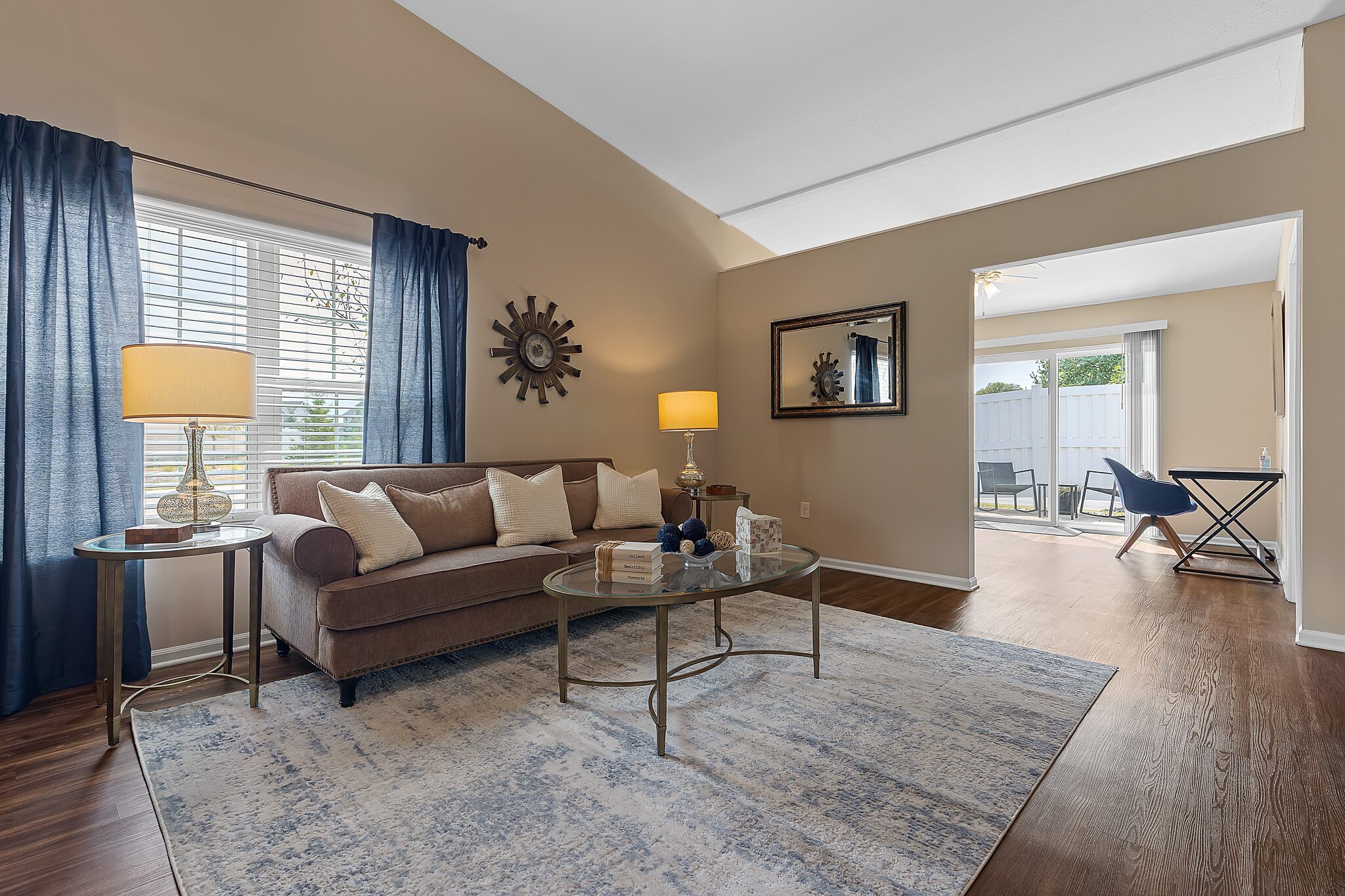 A living room with a brown sofa, glass coffee table, blue and white rug, two table lamps, blue curtains, and a large window, opening to a dining area with patio access.