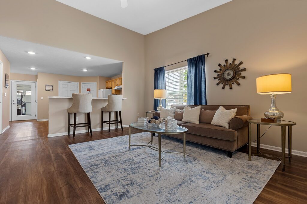 A living room with a brown sofa, glass coffee table, blue-gray rug, and two bar stools at a counter. A lamp, wall clock, and blue curtains are near a window. The floor is wood.