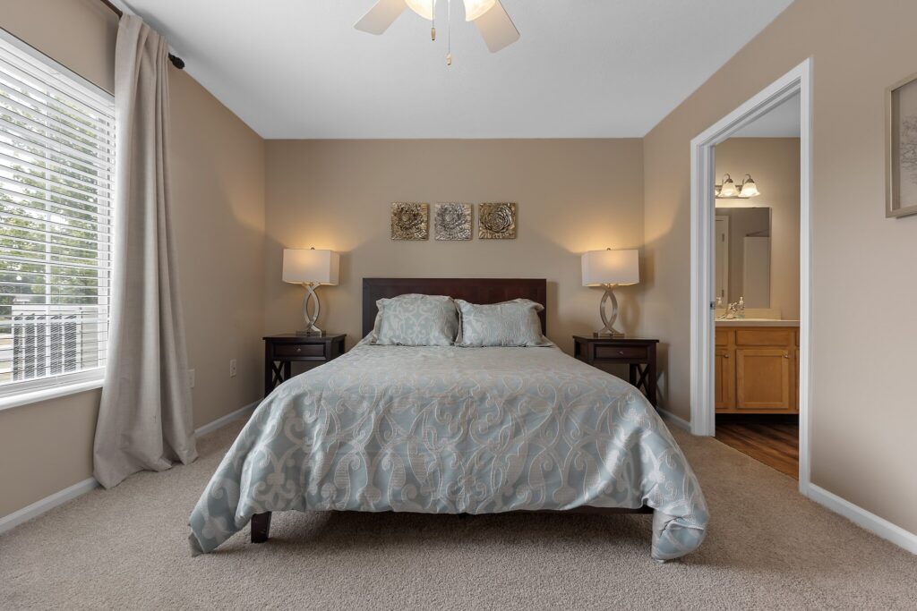 A neatly made bed with two pillows and a patterned comforter sits between two bedside tables with lamps in a softly lit bedroom; a bathroom is visible through an open door.