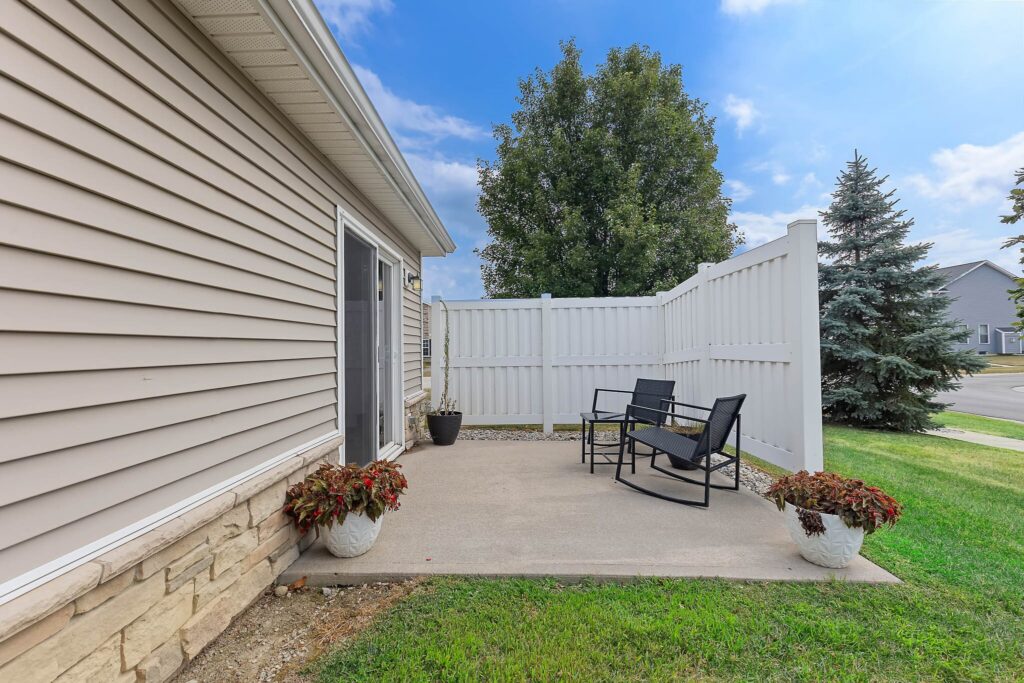 Small concrete patio with two black chairs and a table, bordered by white vinyl fencing, potted plants, and grass, next to a beige house.