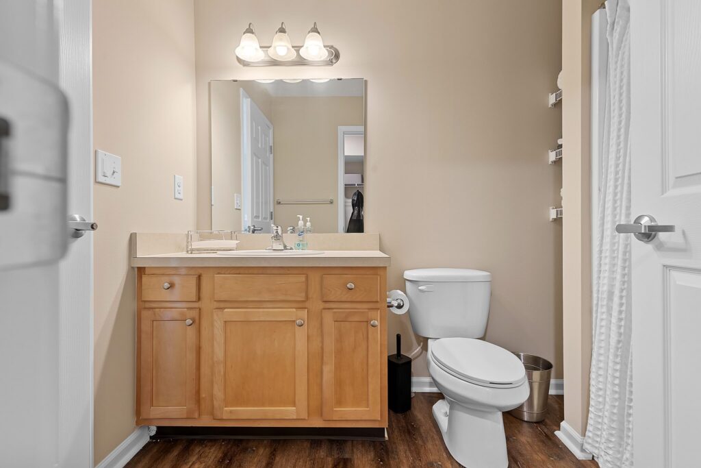 A bathroom with a wooden vanity, large mirror, three-light fixture, toilet, toilet brush, metal trash can, and towel on a wall rack. The floor is wood-style.