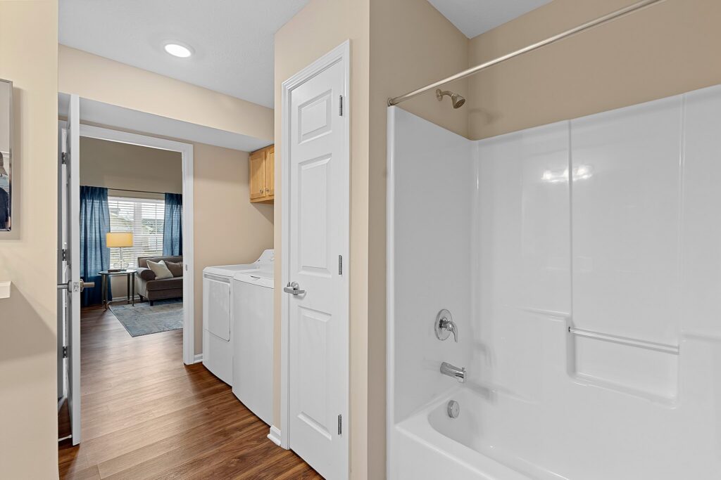 Modern bathroom with a white bathtub and shower combo next to a laundry area with a washer and dryer; living room visible in the background.