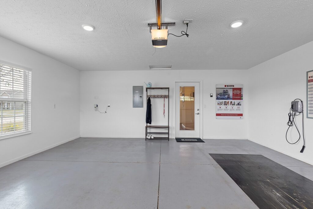 A clean, empty garage with white walls, a concrete floor, a window, a door, a coat rack, an electrical panel, and a wall-mounted device.