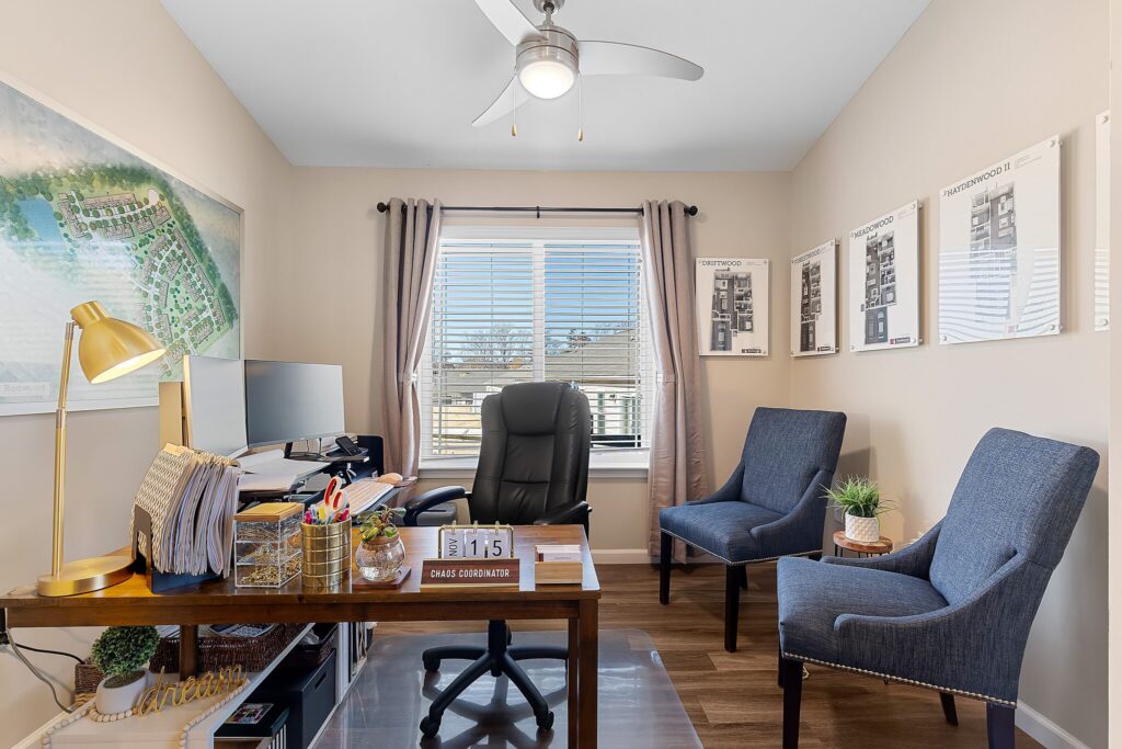A modern home office with a desk, computer, swivel chair, two blue armchairs, wall art, and a window with curtains letting in natural light.