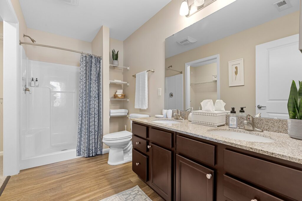 Modern bathroom with a granite double-sink vanity, dark wood cabinets, a toilet, shower with patterned curtain, open shelving, and neutral walls.