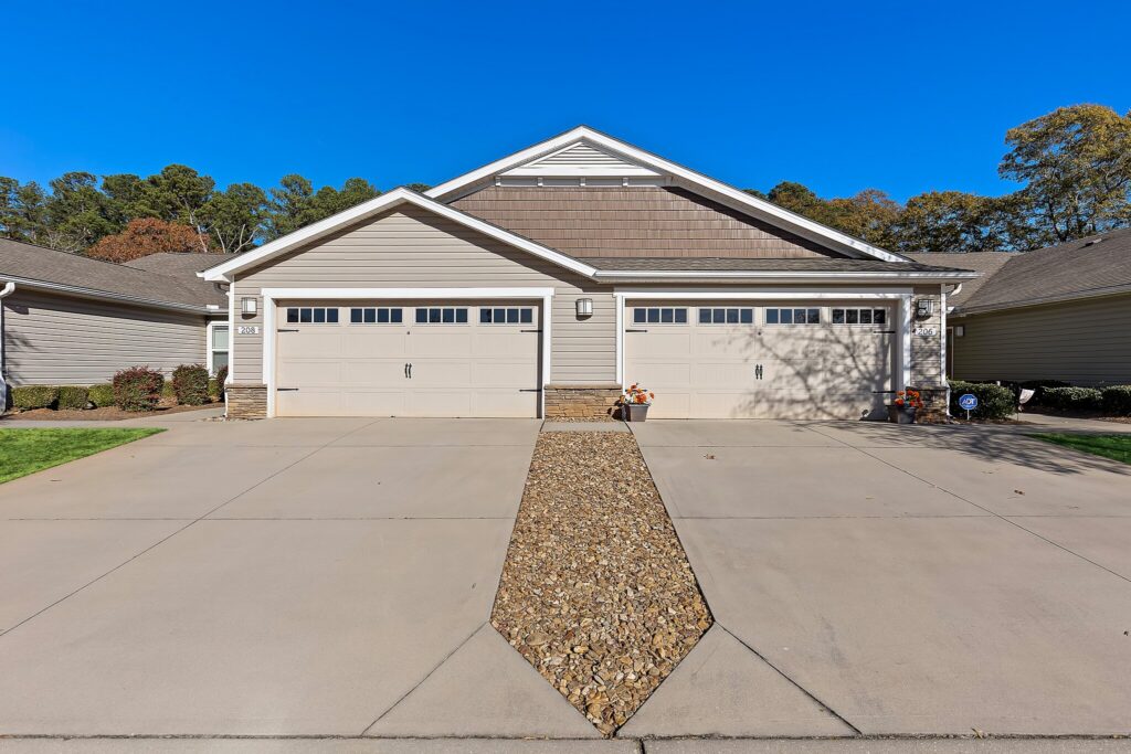 Front view of a duplex with two white double garages, beige siding, gabled roof, stone accents, and a rock-lined median between two concrete driveways.