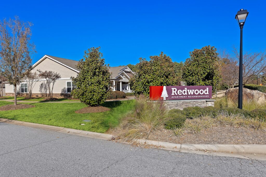 A residential building with a sign in front reading "Redwood Apartment Neighborhoods," surrounded by trees and landscaping under a clear blue sky.