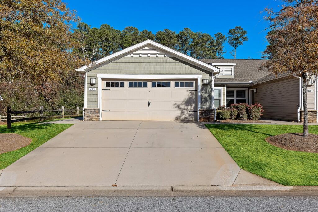 Single-story house with gray siding, attached two-car garage, concrete driveway, and front lawn, with trees and blue sky in the background.