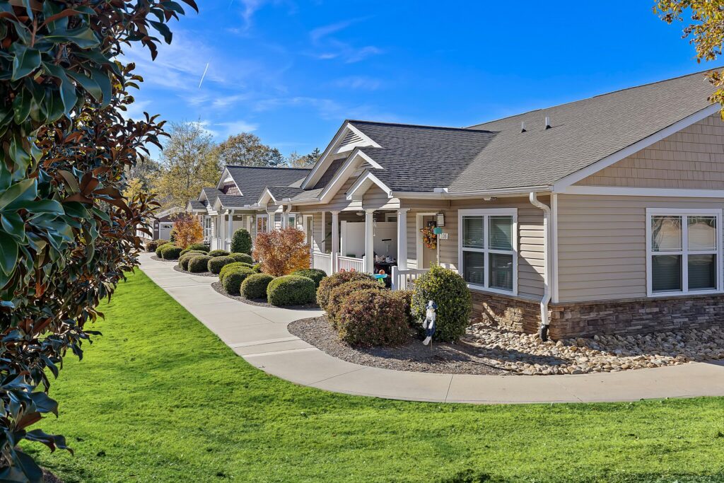 Single-story residential units with beige siding and porches line a curved walkway, bordered by manicured lawns and trimmed shrubs, under a clear blue sky.