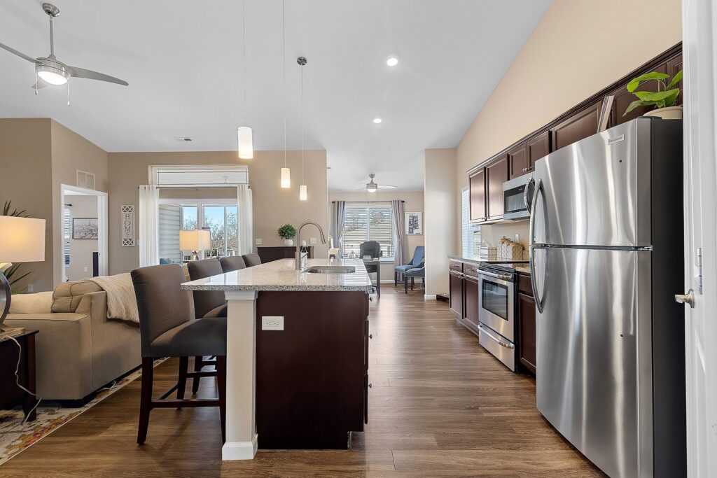 Modern open-concept kitchen and living area with stainless steel appliances, dark wood cabinets, island seating, and large windows letting in natural light.
