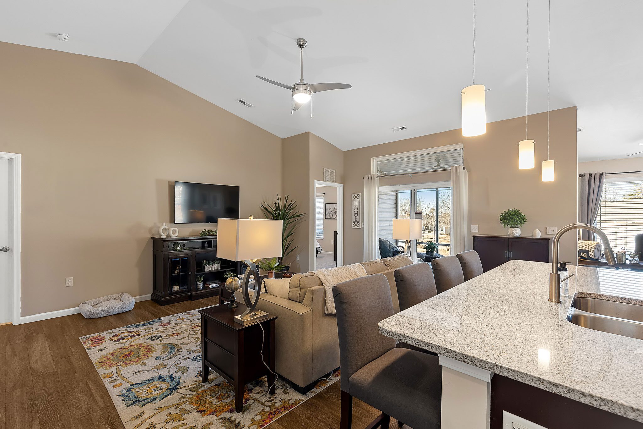 Open-concept living area with kitchen island, pendant lighting, beige walls, brown sofa, TV, and sliding glass doors leading to a patio.