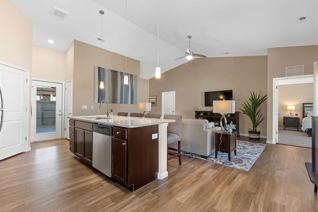 Open-concept living area with a kitchen island, stainless steel appliances, light brown walls, wood flooring, and a living room with a TV and seating area. Bedroom visible through doorway.