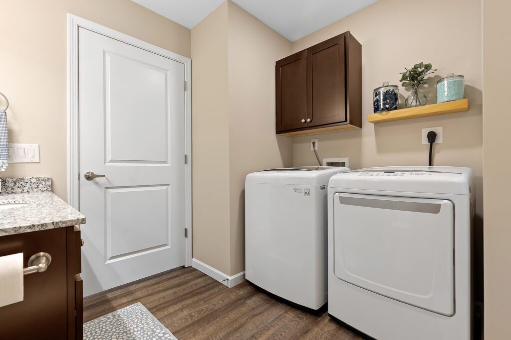 Laundry room with a washing machine and dryer side by side, brown cabinets above, a shelf with plants and decor, and beige walls with wood flooring.