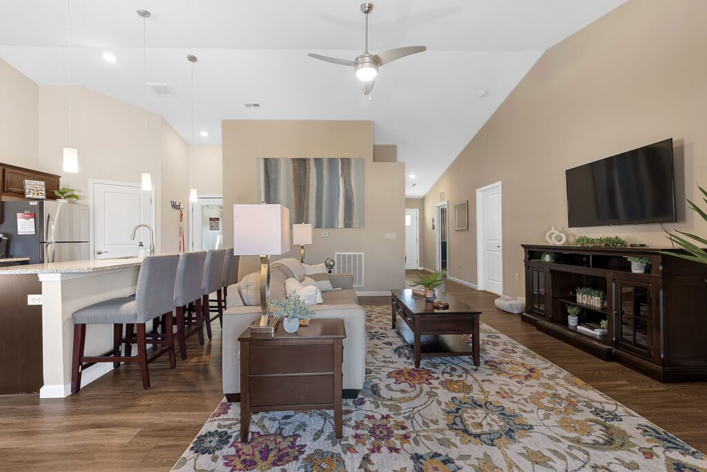 Modern open-concept living room with neutral tones, a patterned area rug, a sofa, a wall-mounted TV, and a kitchen with barstools and pendant lighting in the background.