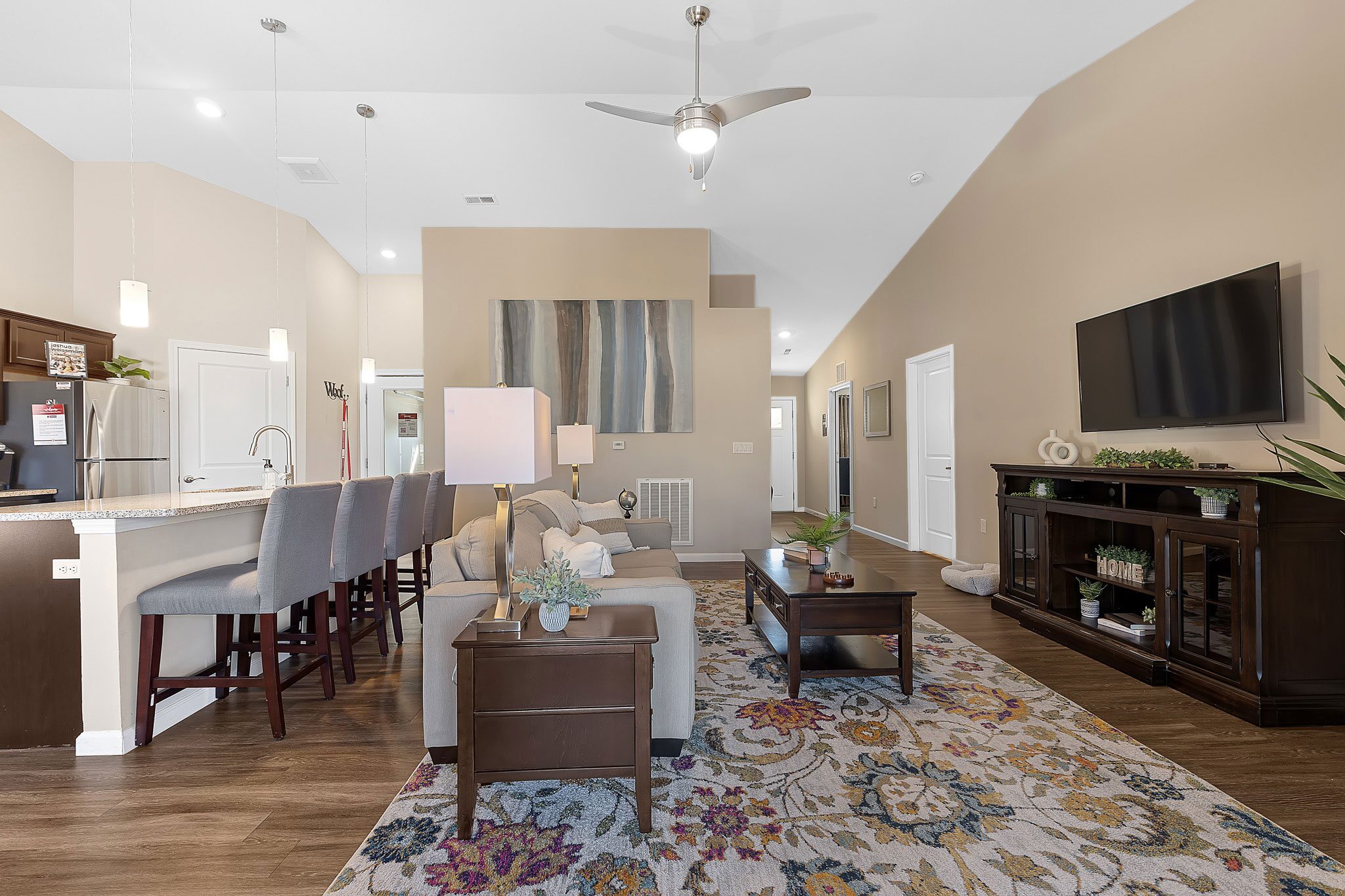 Modern open-concept living room with neutral tones, a patterned area rug, a sofa, a wall-mounted TV, and a kitchen with barstools and pendant lighting in the background.