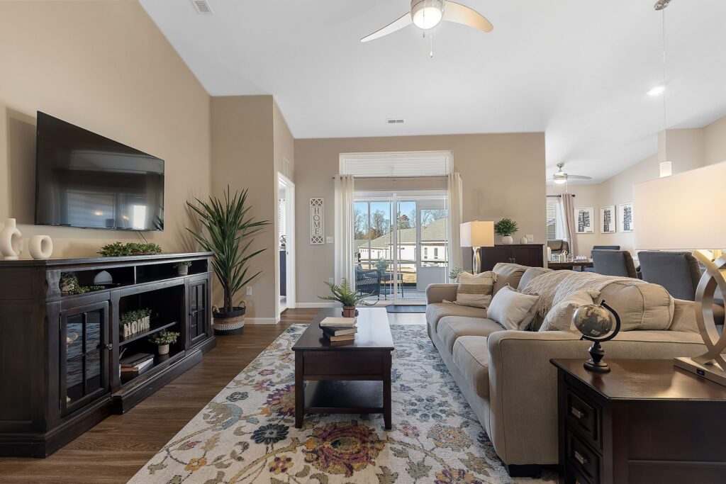 A modern living room with a beige sofa, patterned rug, wall-mounted TV, dark wood furniture, indoor plants, and large windows letting in natural light.
