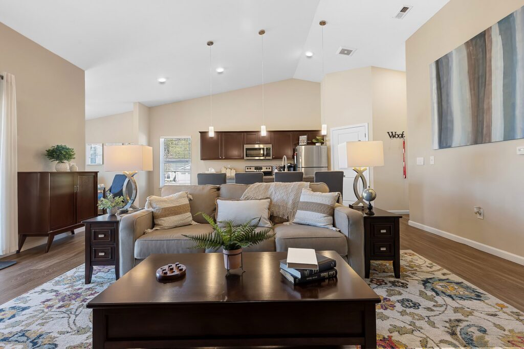Modern living room with a gray sofa, coffee table, and side tables with lamps; open kitchen area in the background with stainless steel appliances.