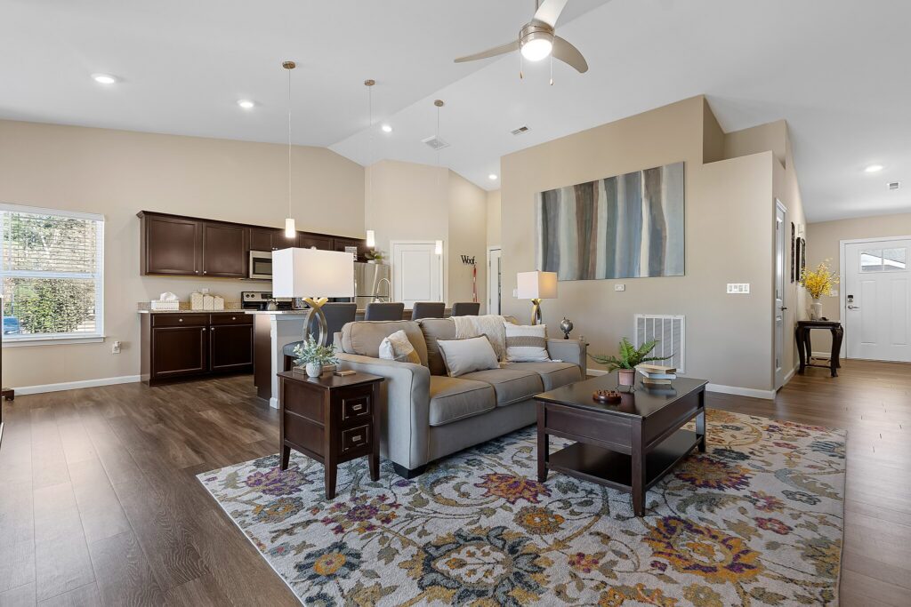 Open-concept living room and kitchen with neutral decor, gray sofa, patterned rug, wooden furniture, stainless steel appliances, and modern lighting.