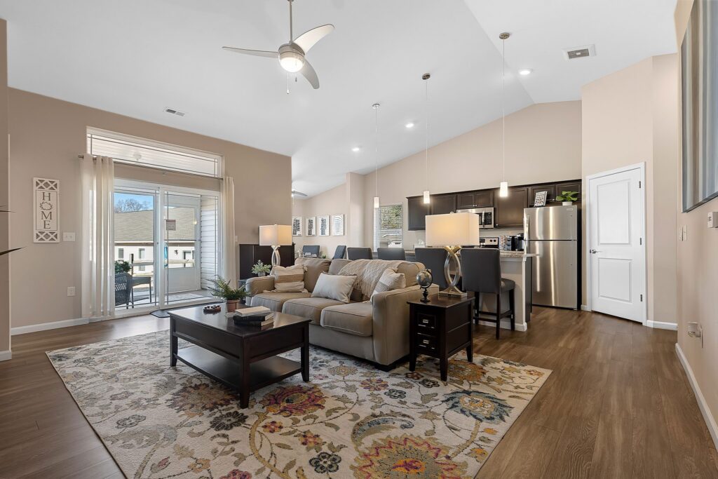 Modern open-concept living room and kitchen with neutral decor, ceiling fan, large windows, and wood flooring. A beige sofa and floral rug are centered in the space.