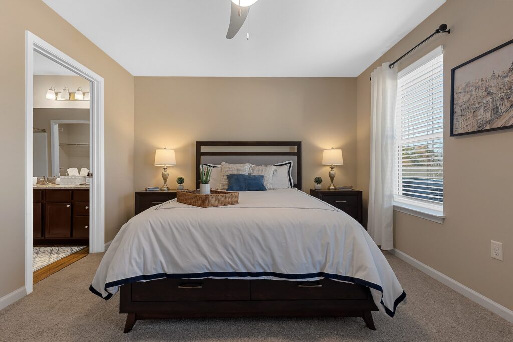 A neatly made bed with white bedding is centered in a beige bedroom, flanked by two dark nightstands with lamps; a window and artwork are on the right wall.