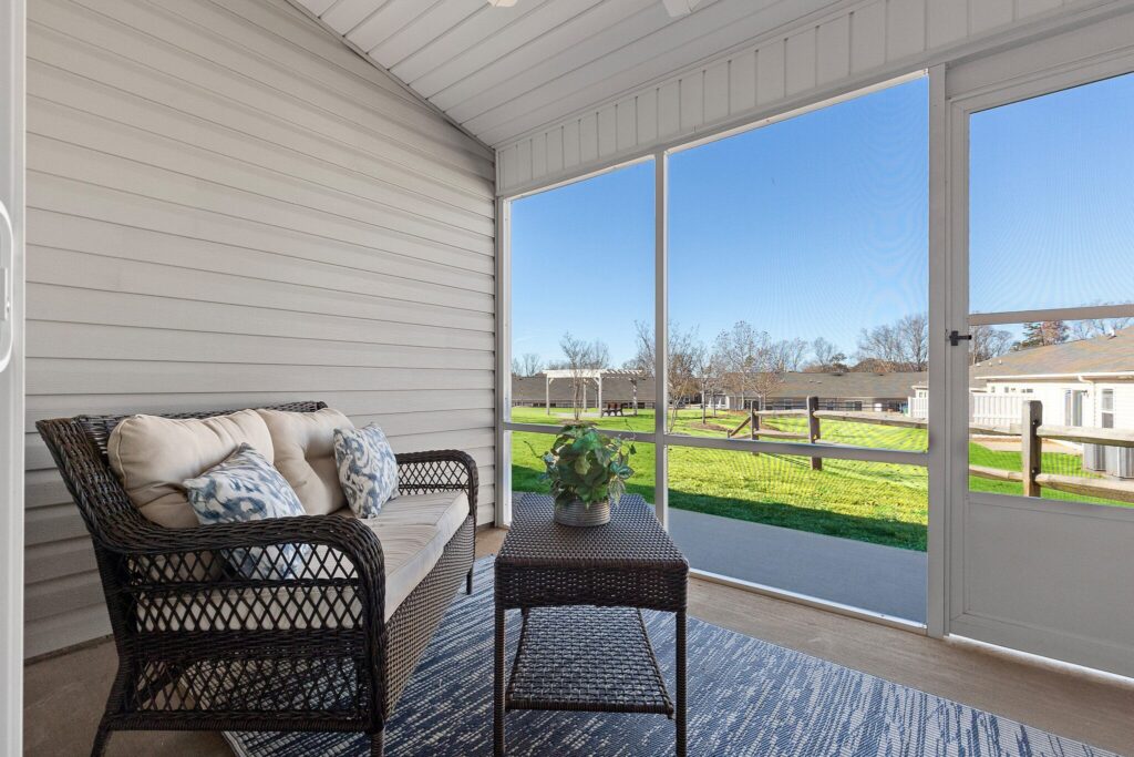 A wicker loveseat and table sit on a rug in a screened-in porch, overlooking a grassy backyard with a fence and clear sky.