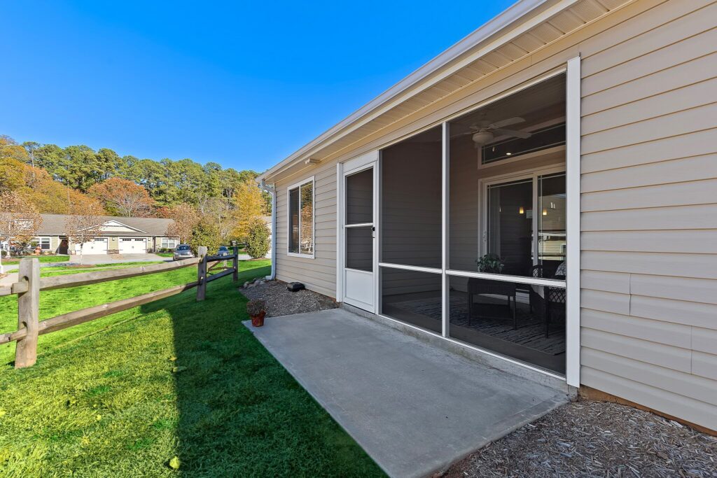 Side view of a beige house with a screened-in porch, concrete patio, green lawn, wooden fence, and trees in the background on a clear day.