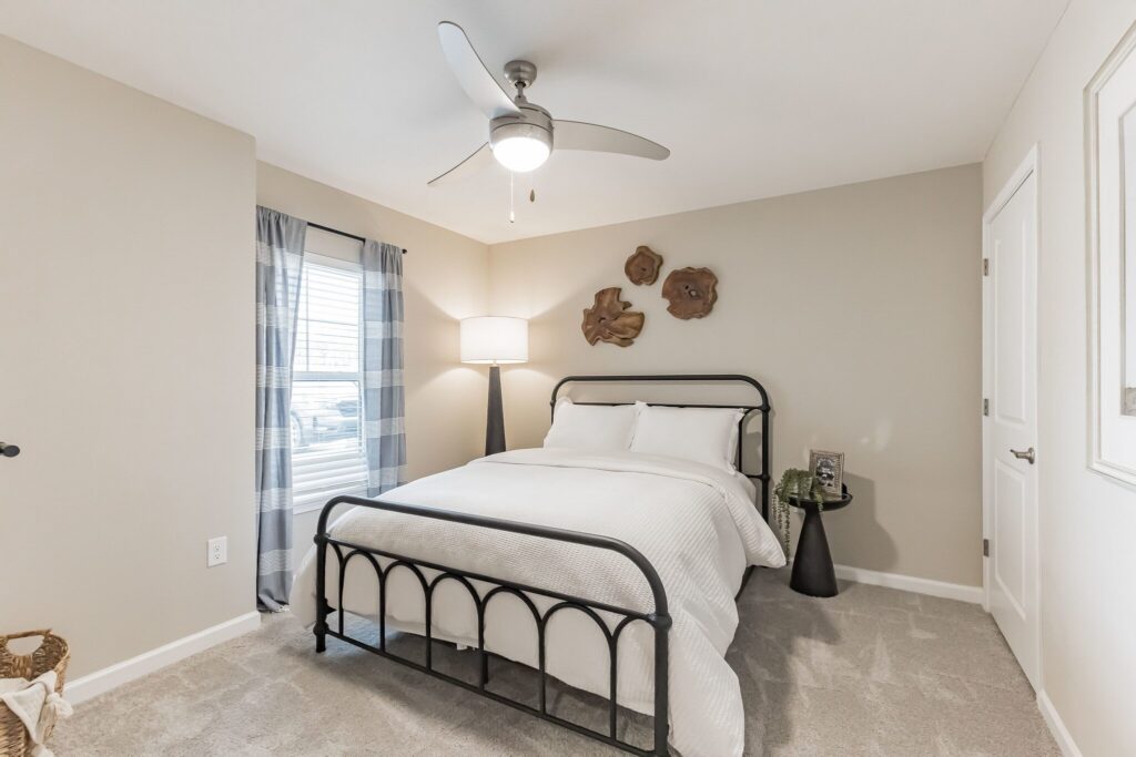 A bedroom with a metal frame bed, white bedding, a floor lamp, a ceiling fan, a side table with a vase, and wall decor above the bed. Light streams in from a window with plaid curtains.