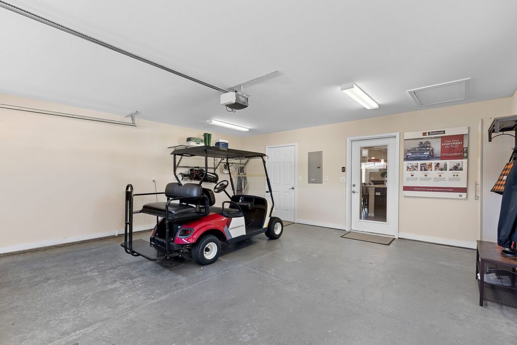 A clean garage with a red and black golf cart parked inside, beige walls, concrete floor, wall-mounted storage, and a door leading to the interior of the house.