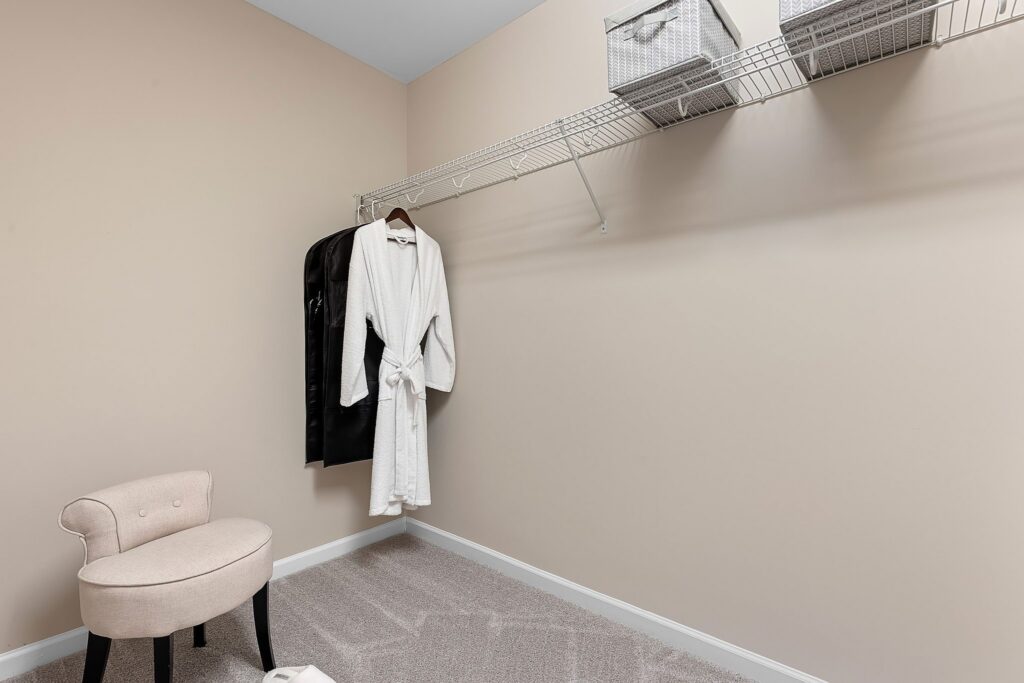 A beige walk-in closet with a wire shelf holding baskets, a white robe and black garment on hangers, and a light upholstered chair on carpeted floor.