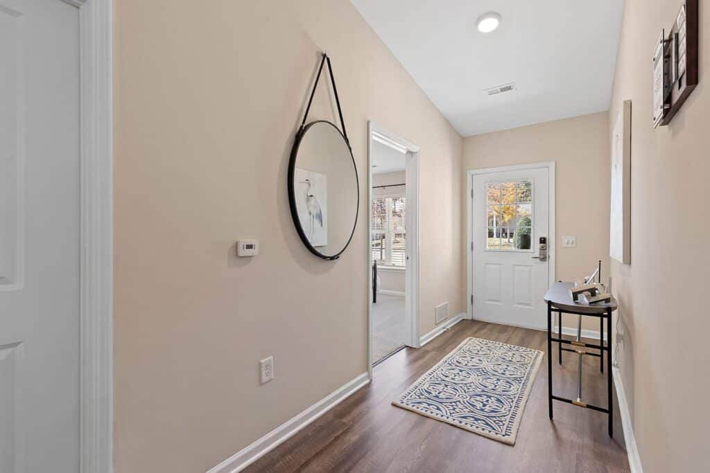 A hallway with beige walls, a round mirror, a small desk with a chair, a rug, and a white door leading outside with windows showing autumn trees.