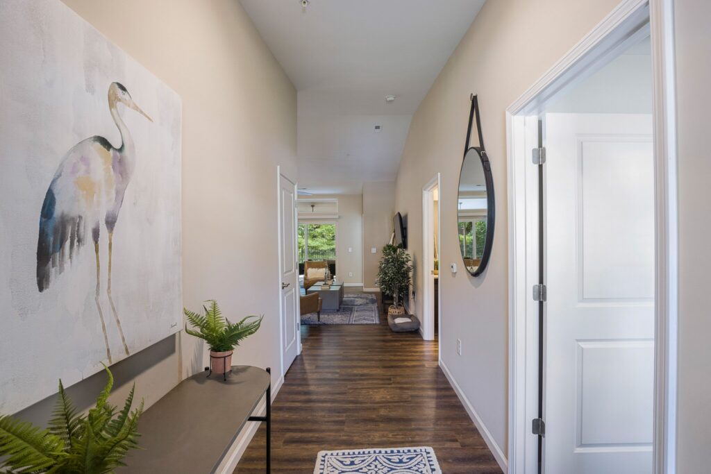 A hallway with wood flooring, a table with potted plants, a bird painting on the wall, and doors leading to a living area with large windows in the background.