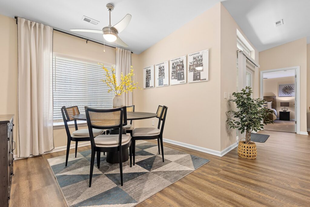 A contemporary dining area with a round table, four chairs, geometric rug, wall art, potted plant, and large window with drapes.