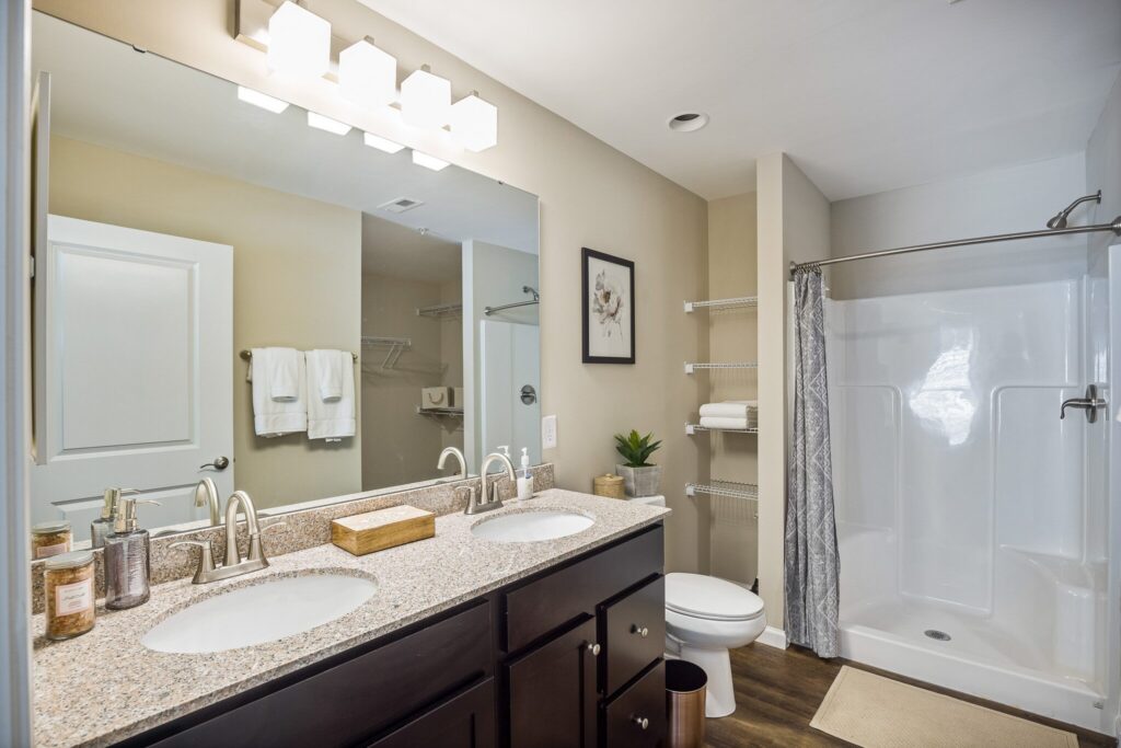 Modern bathroom with double sink vanity, granite countertop, large mirror, walk-in shower, toilet, towel racks, and neutral decor.